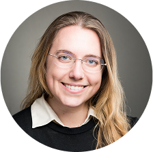 Smiling woman with long blonde hair and glasses, wearing a black sweater over a white collared shirt against a neutral background